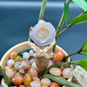 Puede incluir: Primer plano de una geoda de ágata de color púrpura claro y marrón con un centro blanco. La geoda está montada en un palo de madera y envuelta con un cordón trenzado beige. La geoda está sentada en una maceta con pequeñas piedras de color naranja y blanco.