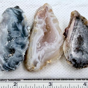 May include: Three polished agate slices in shades of grey, white, and brown. The stones have a natural, organic shape and visible banding patterns. The agate slices are displayed on a white surface next to a ruler.