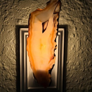 May include: A wall-mounted night light featuring a translucent, irregularly shaped slice of orange and white agate stone. The stone is backlit, revealing natural banding and crystalline textures, mounted on a rectangular white plastic base.