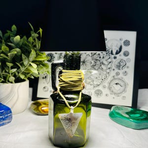 May include: A unique lamp made from a green glass bottle, featuring a black lampshade. The bottle is adorned with a leather wrap and a triangular pendant. The background includes a framed drawing and decorative stones.