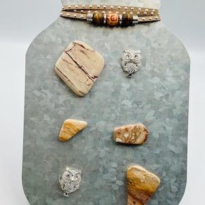 Puede incluir: Un adorno de pared decorativo con forma de tarro de metal con una correa de cuero y cuentas de madera. El tarro presenta varias piedras naturales y dos dijes de búho plateados. Las piedras son en tonos marrón, beige y crema.