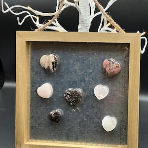 May include: A wooden frame with a gray background holds seven heart-shaped stones. The stones are in various shades of pink, white, and black.