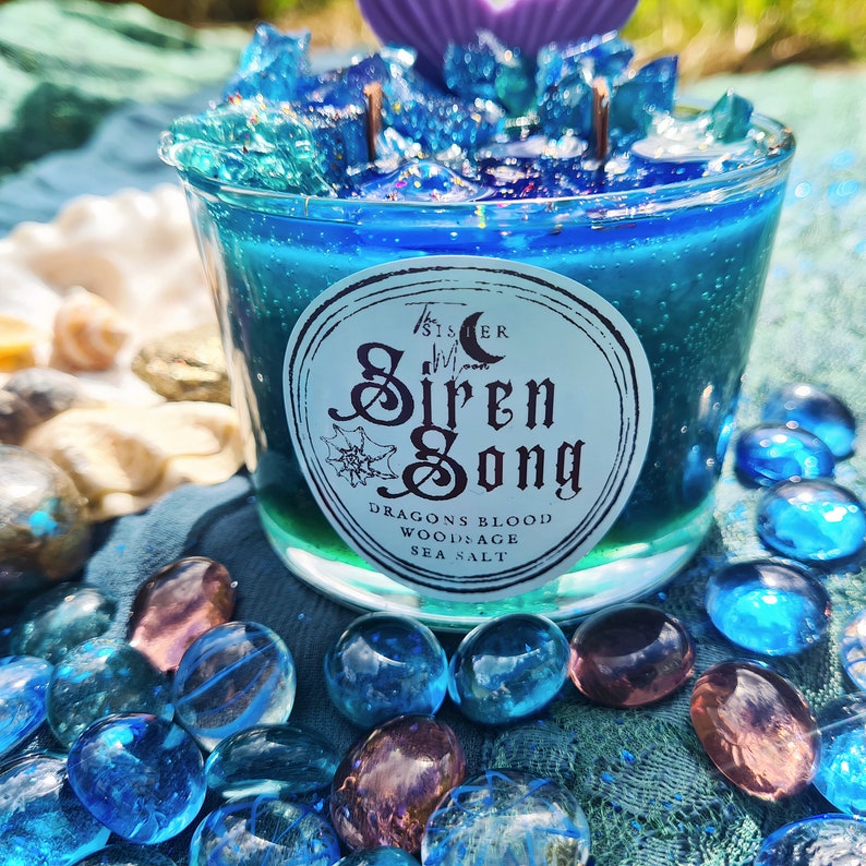 May include: A blue and green glass candle jar with a white label that reads "The Sister Moon Siren Song Dragons Blood Woodsage Sea Salt". The candle is surrounded by blue and purple glass stones.