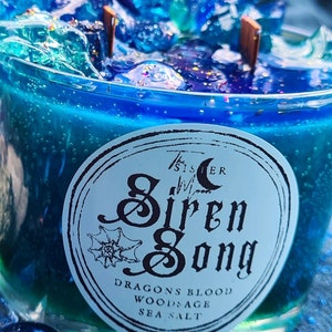 May include: A blue glass candle with a mermaid tail on top. The candle is labeled "The Sister Moon Siren Song" and has the ingredients "Dragons Blood, Wood Sage, Sea Salt" listed below the label.