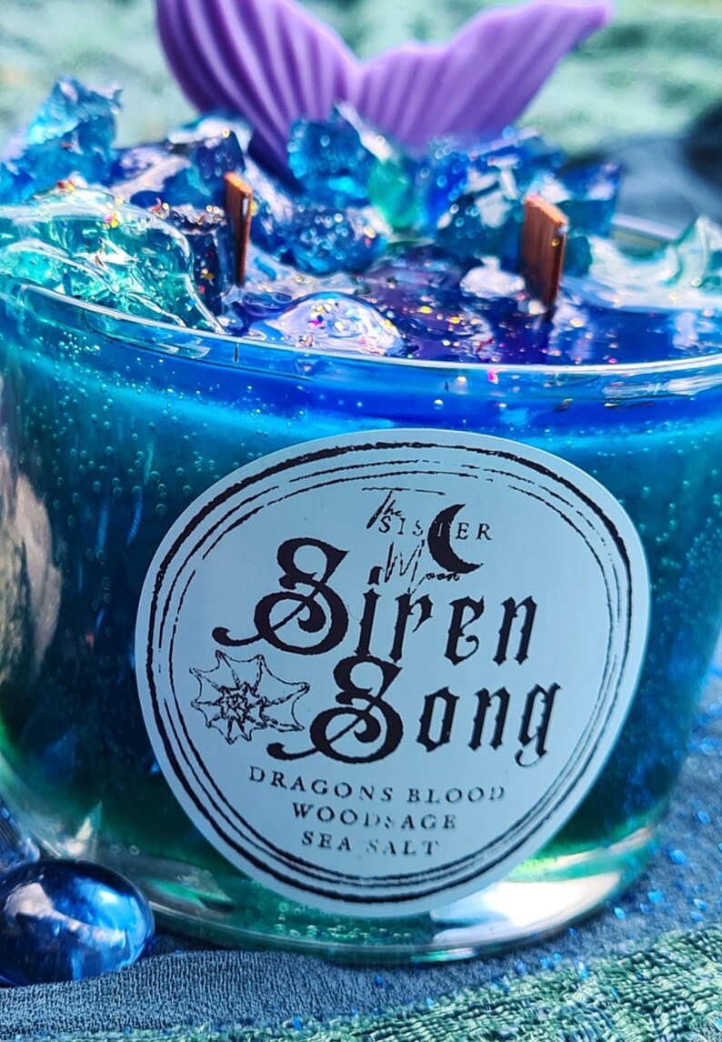 May include: A blue glass candle with a mermaid tail on top. The candle is labeled "The Sister Moon Siren Song" and has the ingredients "Dragons Blood, Wood Sage, Sea Salt" listed below the label.