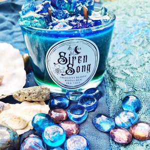May include: A blue glass candle jar with a white label that reads "The Sister Siren Song Dragons Blood Woodsage Sea Salt". The candle is surrounded by blue and iridescent glass stones.