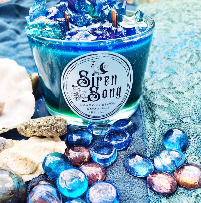 May include: A blue glass candle jar with a white label that reads "The Sister Siren Song Dragons Blood Woodsage Sea Salt". The candle is surrounded by blue and iridescent glass stones.