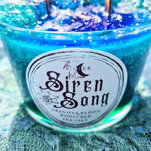 May include: A teal blue candle with a white label that reads "The Sister Moon Siren Song Dragons Blood, Woodsage, Sea Salt".