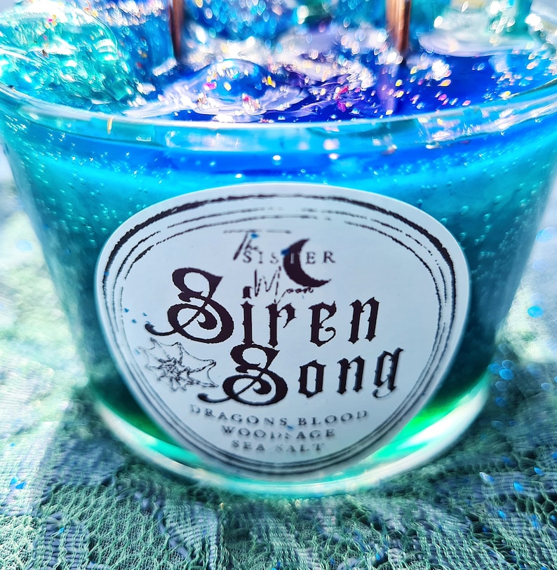 May include: A teal blue candle with a white label that reads "The Sister Moon Siren Song Dragons Blood, Woodsage, Sea Salt".