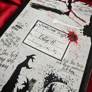Puede incluir: Una cubierta de cuaderno con una estética grunge y desgastada. La superficie blanca presenta salpicaduras de tinta negra, una calavera y siluetas de manos con garras. El texto incluye "DO NOT TOUCH" y "Hawkins High, Billy H."
