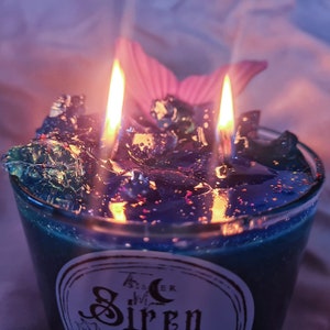 May include: A teal blue glass candle with a black and silver glitter top. The candle has two wicks and is lit. The candle is labeled "Tisser  Siren".