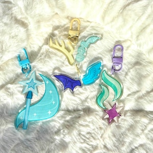 May include: A set of six colorful acrylic keychains featuring various whimsical designs, including a crescent moon with a star, a bat, wings, a flame, a star, and antlers. Each keychain has a metal ring and a clear acrylic charm.
