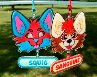 Op de afbeelding: Twee kleurrijke cartoon dierenkop sleutelhangers met de namen "Squiq" en "Sanguine" aan de onderkant. De sleutelhanger aan de linkerkant is blauw en rood met een roze neus en witte sterren in de ogen. De sleutelhanger aan de rechterkant is oranje en rood met een witte neus en zwarte ogen.
