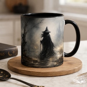 May include: A ceramic mug with a black handle and interior, featuring a printed illustration of a cloaked figure in a pointed hat standing in a misty, dark forest landscape. The scene includes bare trees and a somber, atmospheric color palette.