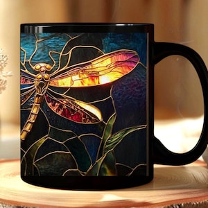 Puede incluir: Una taza de cerámica negra con un gráfico estilo vidriera de una libélula. La libélula tiene segmentos corporales dorados y alas con tonos iridiscentes de naranja, amarillo y rojo sobre un fondo azul profundo y verde oscuro.
