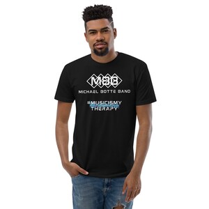 Könnte beinhalten: Schwarzes T-Shirt mit dem weißen Schriftzug "MICHAEL BOTTE BAND" über dem Hashtag "#MUSICISMY THERAPY" in Blau und Weiß. Das Shirt hat ein weißes geometrisches Logo. Die Person trägt eine blaue Jeans.