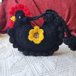 May include: A black crochet chicken-shaped bag with a red comb and yellow accents. The bag features a yellow flower and a black strap. The bag is sitting on a white quilted surface.