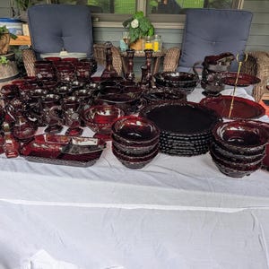 May include: A large collection of dark red pressed glass tableware, including numerous goblets, bowls, plates, a pitcher, and a tiered serving stand. The glassware features intricate geometric and floral patterns, with a deep ruby hue throughout the set.