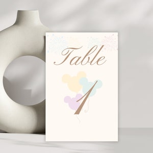 Puede incluir: Número de mesa uno con fondo blanco y texto marrón. El texto dice "Table" y el número "1" está en forma de cabeza de Mickey Mouse marrón. Hay tres globos con forma de cabeza de Mickey Mouse en el fondo, uno amarillo, uno azul y uno morado.