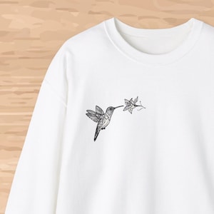 May include: White crewneck sweatshirt with a black and white hummingbird graphic on the left chest. The hummingbird is flying towards a flower.