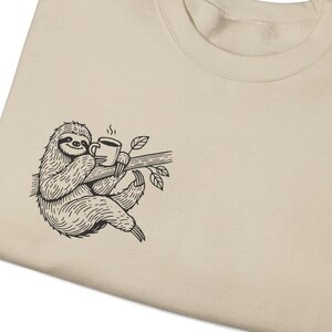 May include: A beige sweatshirt with a black and white illustration of a sloth holding a cup of coffee on a branch.