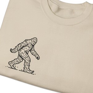 May include: A beige t-shirt with a black line drawing of a Bigfoot walking on the left side of the shirt.