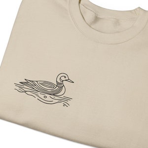 May include: A beige sweatshirt with a black line drawing of a duck swimming on the front.