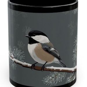 May include: A black coffee mug with a gray background featuring a black-capped chickadee perched on a snow-covered branch. The bird has a black head, white cheeks, and a gray body.
