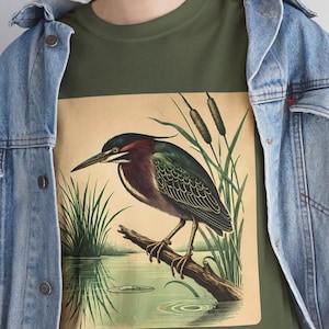 May include: A green t-shirt with a vintage illustration of a green heron perched on a branch over a pond. The heron has a long, thin beak and is surrounded by reeds and cattails.