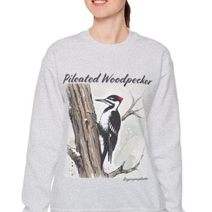May include: A light gray sweatshirt featuring a watercolor illustration of a Pileated Woodpecker perched on a tree branch. The text "Pileated Woodpecker" is above the bird. The bird is black and white with a red crest.