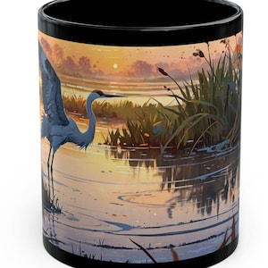 May include: A black ceramic mug with a colorful image of a blue heron standing in a shallow body of water at sunset. The water reflects the orange and pink hues of the sky. Green reeds and grasses line the edge of the water.