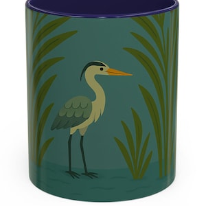 May include: A ceramic mug with a teal background featuring a cartoon heron illustration. The heron is gray and tan with a yellow beak, standing in a marsh with green reeds. The interior of the mug is a deep blue.
