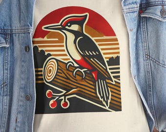 May include: Cream-colored t-shirt featuring a stylized woodpecker perched on a log. The design includes a red, black, and white woodpecker, a brown log, and a sunset-style background. The shirt is worn by a person wearing a denim jacket.