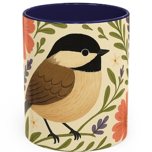 May include: A ceramic coffee mug with a navy blue interior and a cream-colored exterior. The mug features a detailed illustration of a bird perched on a branch, surrounded by flowers and foliage. The bird has a black head, brown body, and beige chest.