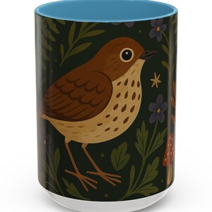 May include: A ceramic coffee mug with a teal interior and a wrap-around design featuring a brown bird with a tan breast, set against a dark green background with floral and mushroom accents. The mug has a white base.