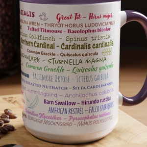 May include: A white ceramic mug with a purple handle. The mug features a list of common North American birds with their scientific names. The text is in pink, purple, and blue. The mug is sitting on a wooden surface with a spoon and a macaron.