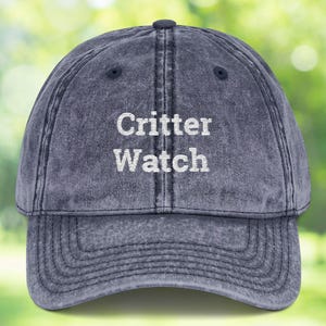 May include: A faded navy blue baseball cap with the words "Critter Watch" embroidered in white. The cap has a curved brim and a classic six-panel design, perfect for outdoor activities.
