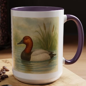 May include: A white ceramic mug with a purple rim and handle. The mug features a watercolor illustration of a red-headed duck swimming in a pond with green reeds.