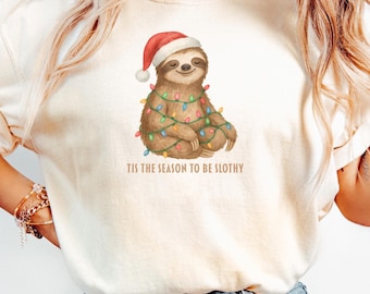 May include: Ivory-colored t-shirt featuring a sloth wearing a Santa hat and wrapped in Christmas lights. The text "TIS THE SEASON TO BE SLOTHY" is printed below the sloth illustration. The shirt is made of a soft material.
