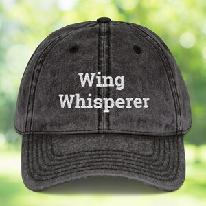 May include: A distressed black baseball cap with the words "Wing Whisperer" embroidered in white. The cap has a curved brim and a classic six-panel design, suitable for casual wear.