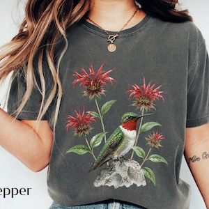 May include: A charcoal gray short-sleeve t-shirt featuring a graphic print of a hummingbird with a red throat perched on a rock, surrounded by red bee balm flowers with green leaves. The word Pepper is printed in black text on the lower left side.