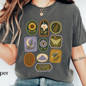May include: A charcoal gray short-sleeve t-shirt featuring a graphic print of various nature-themed icons arranged in a grid. The designs include a sunflower, a crescent moon, a moth, a fern, a flower with an eye, and a cloud, all in muted tones.