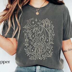 May include: A charcoal grey short-sleeved t-shirt featuring a cream-coloured line art graphic of a winged gargoyle perched on a pedestal. The design includes a crescent moon, stars, a hanging lantern, a key, and gothic architectural elements.