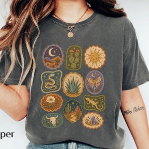 May include: A charcoal gray t-shirt featuring a grid of twelve distinct illustrations including a crescent moon, cactus, sun, snake, agave, moth, flower, desert landscape, lizard, and a skull, all arranged in a rectangular pattern on the chest.