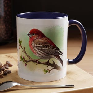 May include: A white ceramic mug with a blue rim and handle features a watercolor illustration of a red and brown bird perched on a branch. The bird has a red head and a brown body with white stripes. The background is a green watercolor wash.