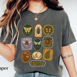May include: A charcoal gray short-sleeve t-shirt featuring a graphic print of various nature-themed illustrations arranged in rectangular and oval frames, including a moth, beetle, butterfly, bee, dragonfly, ladybug, flowers, moon, and fern.
