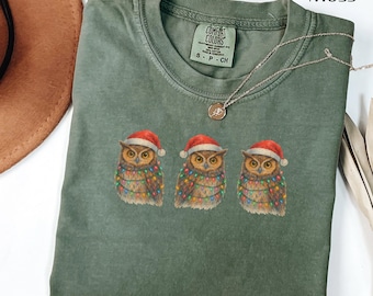 May include: A moss green t-shirt featuring three owls wearing Santa hats and wrapped in Christmas lights. The shirt has a crew neck and a small tag that says "Comfort Colors". A silver necklace with a small pendant is draped across the shirt.