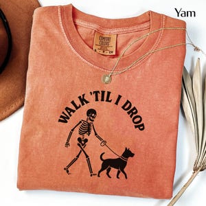 May include: Orange Comfort Colors t-shirt with the phrase "WALK 'TIL I DROP" and a skeleton walking a dog. A gold necklace with a pendant is on the shirt. The shirt is folded. A brown hat and dried plant are in the background.