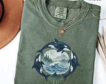 May include: Moss green t-shirt with a circular design of three orcas encircling a seascape with mountains, waves, and a crescent moon. The shirt has a crew neck and a Comfort Colors tag. A silver necklace with a pendant is draped on the shirt.