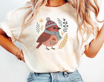 May include: Ivory-colored t-shirt featuring a whimsical illustration of a bird wearing a winter hat and scarf, surrounded by floral and leaf accents. The bird is depicted in shades of brown and pink, with a blue and gray scarf. The word "Ivory" is printed at the bottom.